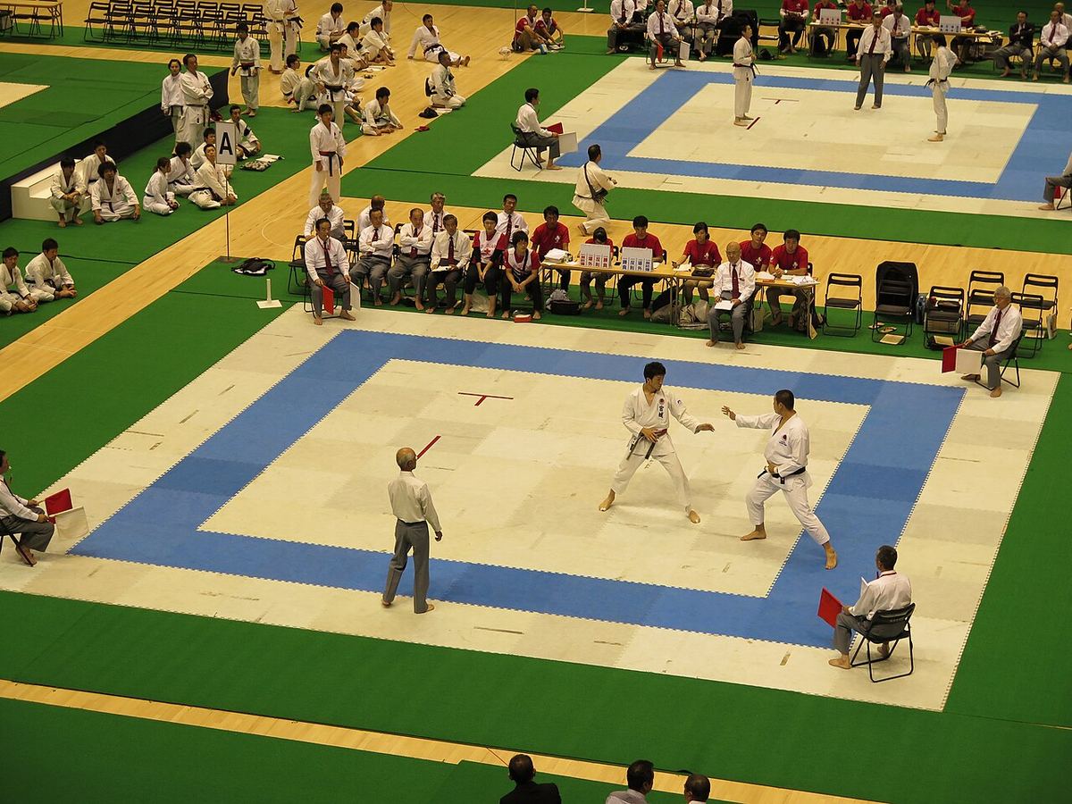 Doi bărbați în echipament de karate se luptă în timpul unei competiții de sparring, într-o sală sportivă.