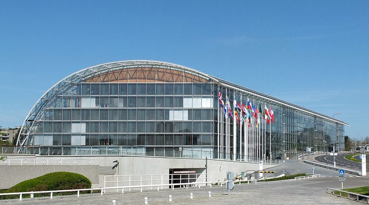 Sediul Băncii Europene de Investiții din Luxemburg, fotografiat din sud, cu steaguri în fața clădirii de sticlă.