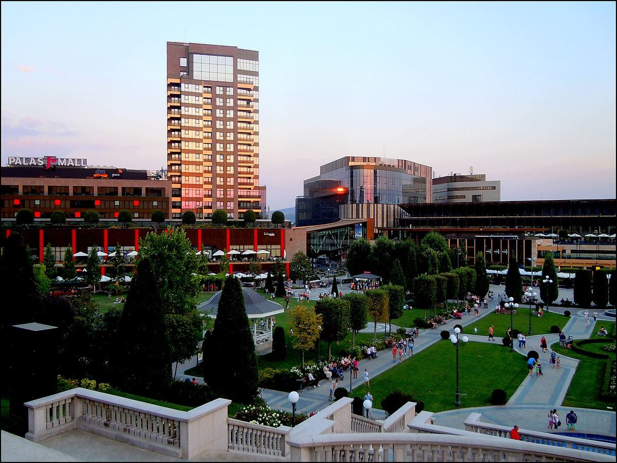 Vedere a clădirii Palas Mall din Iași, cu grădina publică din față și un turn de birouri în fundal, la apus.