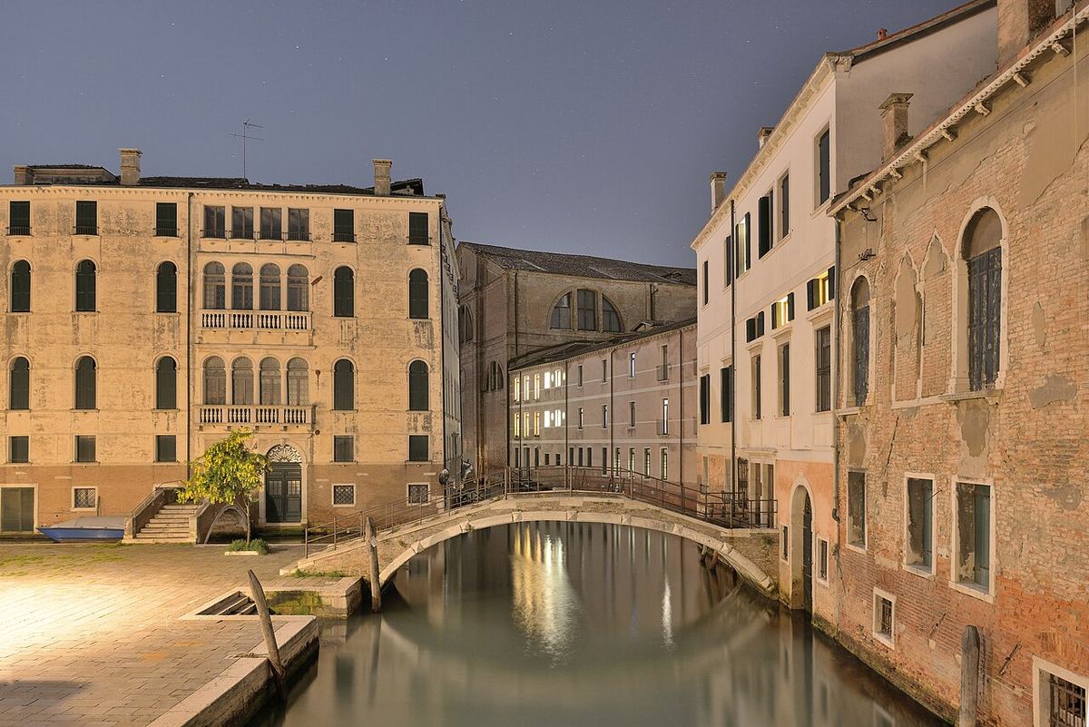 Canal îngust din Veneția noaptea, cu un pod peste apă și clădiri istorice de cărămidă și tencuială.