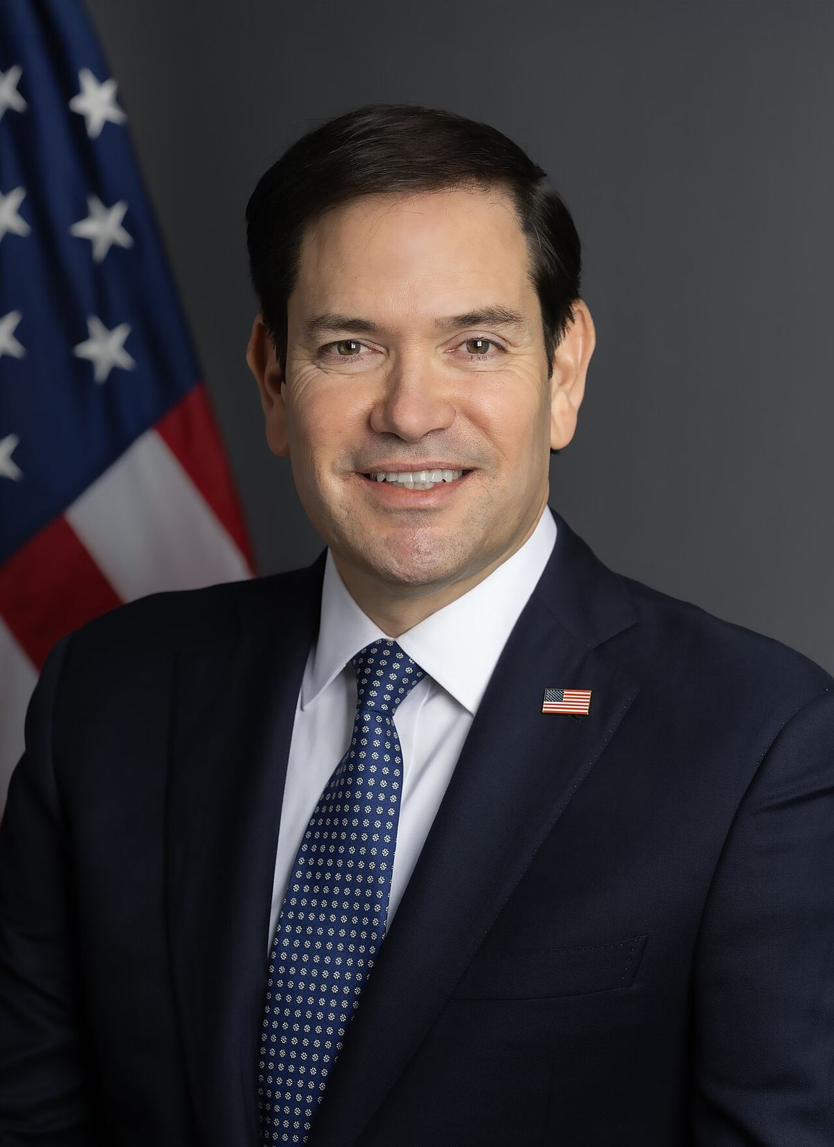 Portretul oficial al secretarului de stat al SUA, Marco Rubio, în costum închis, în fața drapelului american.