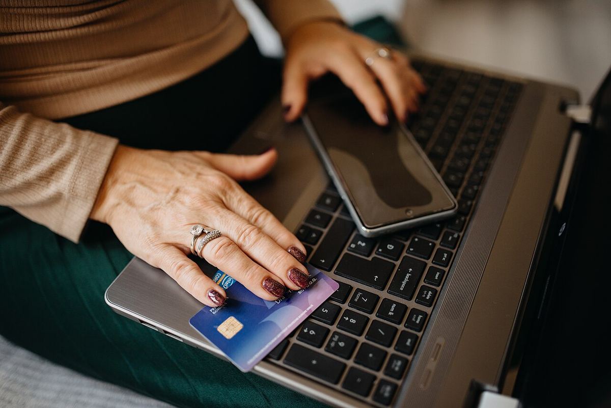 Mâini de femeie ținând un card bancar albastru și un smartphone deasupra unei tastaturi de laptop, ilustrând plăți online.