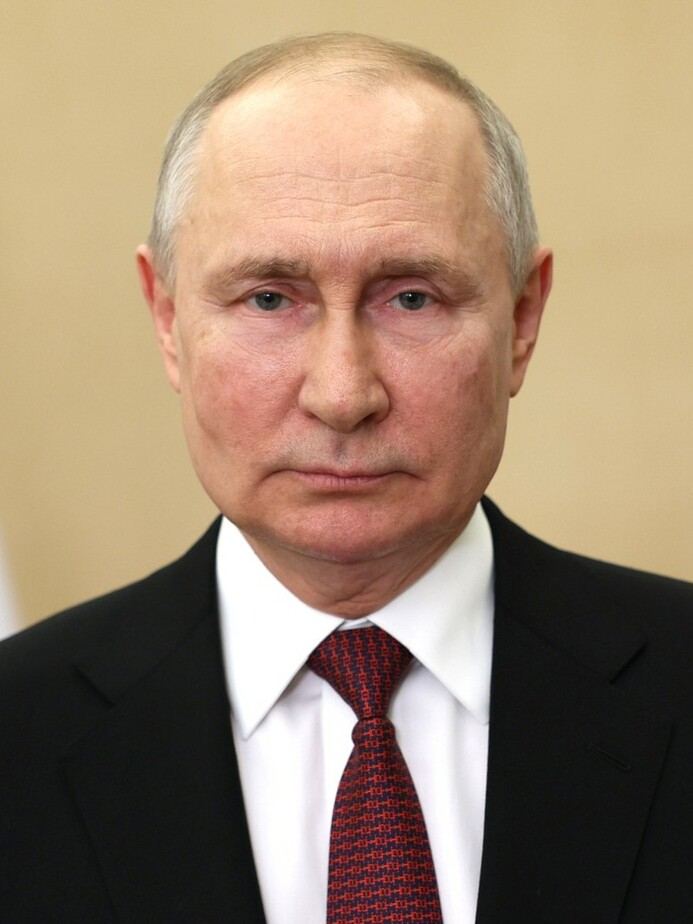 Fotografie de arhivă cu Vladimir Putin, în costum închis, cămașă albă și cravată roșie, pe fundal bej.