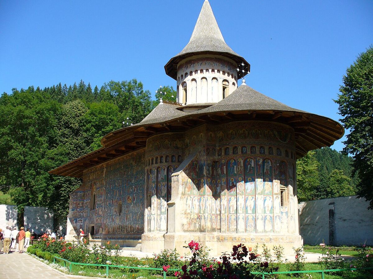 Mănăstirea Voroneț din Bucovina, cu pereții exteriori pictați cu fresce și copaci verzi în jur, pe o zi însorită.
