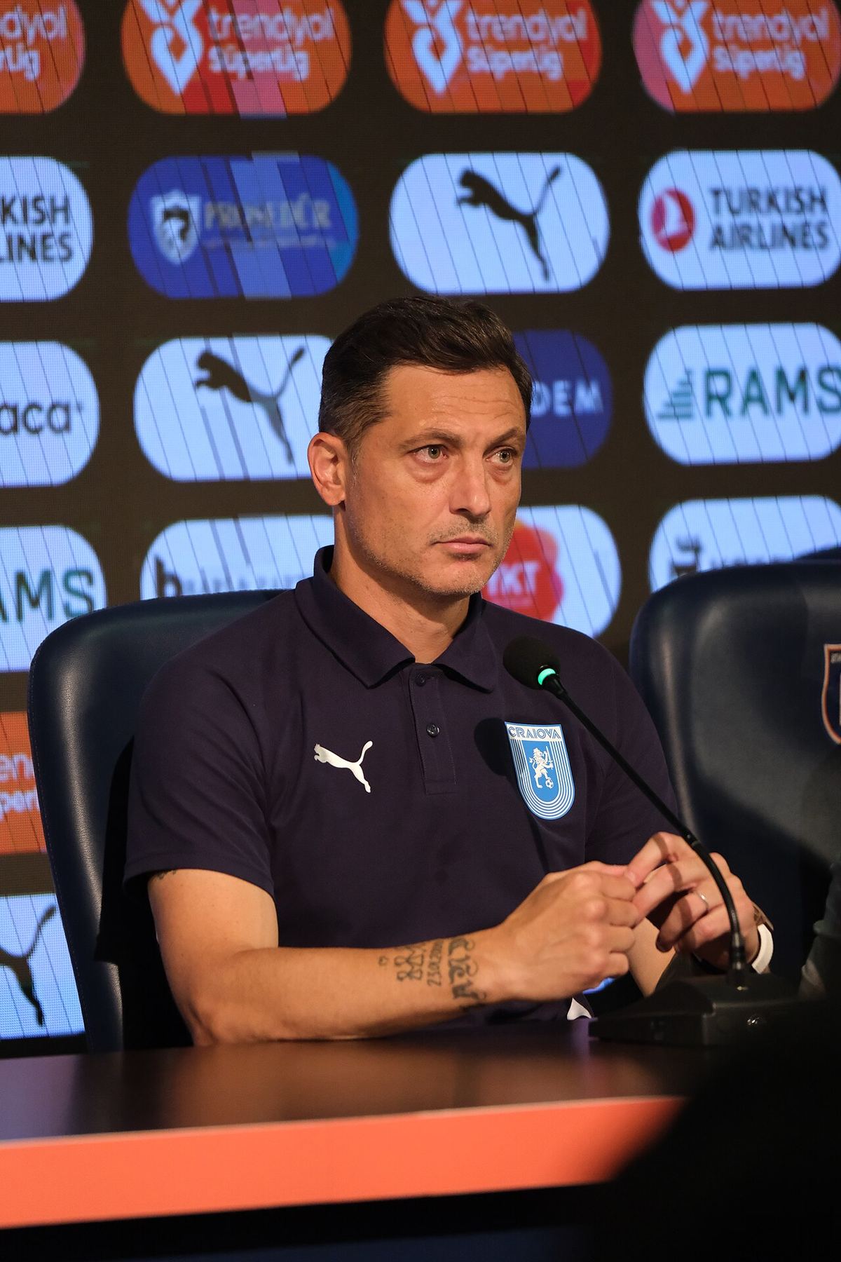 Mirel Rădoi, în tricou polo bleumarin, la o conferință de presă, cu microfonul în față și mâinile împreunate pe masă.