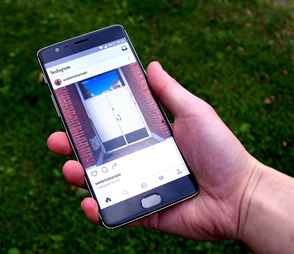 Mână ținând un smartphone cu aplicația Instagram deschisă, pe un fundal de iarbă verde.