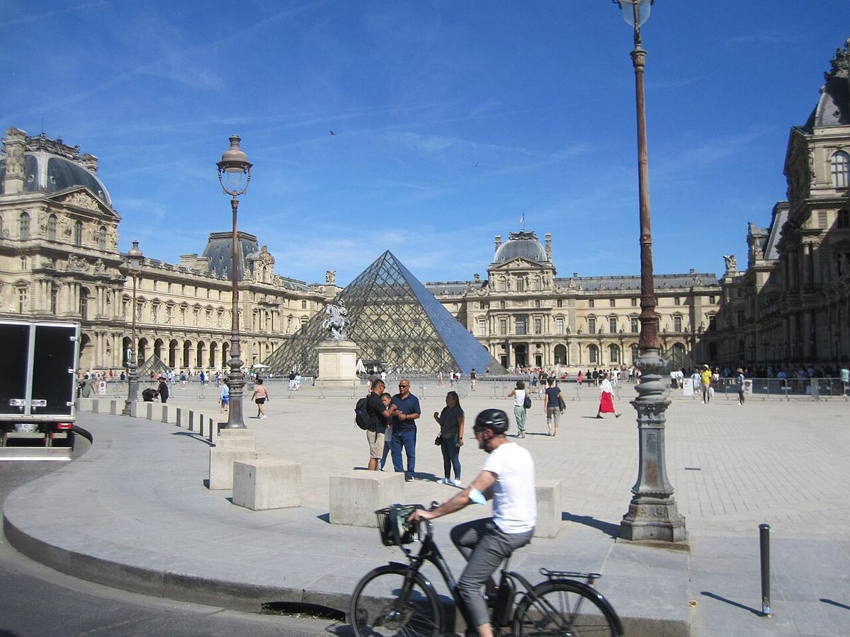 Muzeul Luvru din Paris și Piramida de sticlă din Cour Napoléon, fotografiate ziua, cu turiști și o bicicletă în prim-plan (fotografie de arhivă).