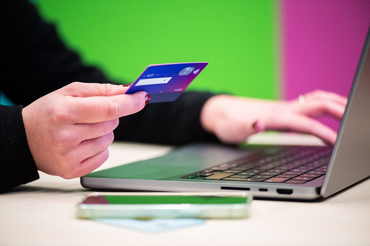 Mână ținând un card bancar albastru, în timp ce o altă persoană tastează pe laptop, pentru cumpărături online.