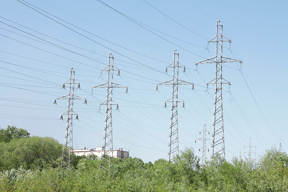 Stâlpi metalici de înaltă tensiune și linii electrice aeriene deasupra unei zone cu vegetație verde, sub cer senin.