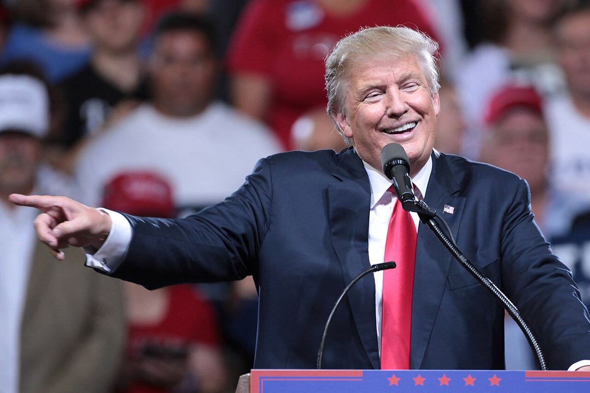 Donald Trump vorbind la microfon la un miting electoral, arătând cu mâna spre public, în fața unei mulțimi de susținători.