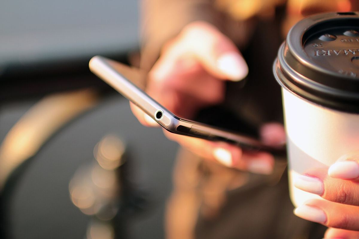 Mână care ține un telefon mobil, lângă o cafea
