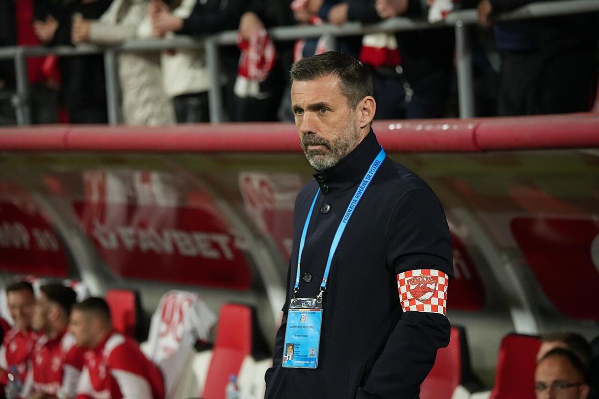 Zeljko Kopic, antrenor de fotbal, cu banderolă de căpitan și acreditare, în fața băncii de rezerve a unui stadion.