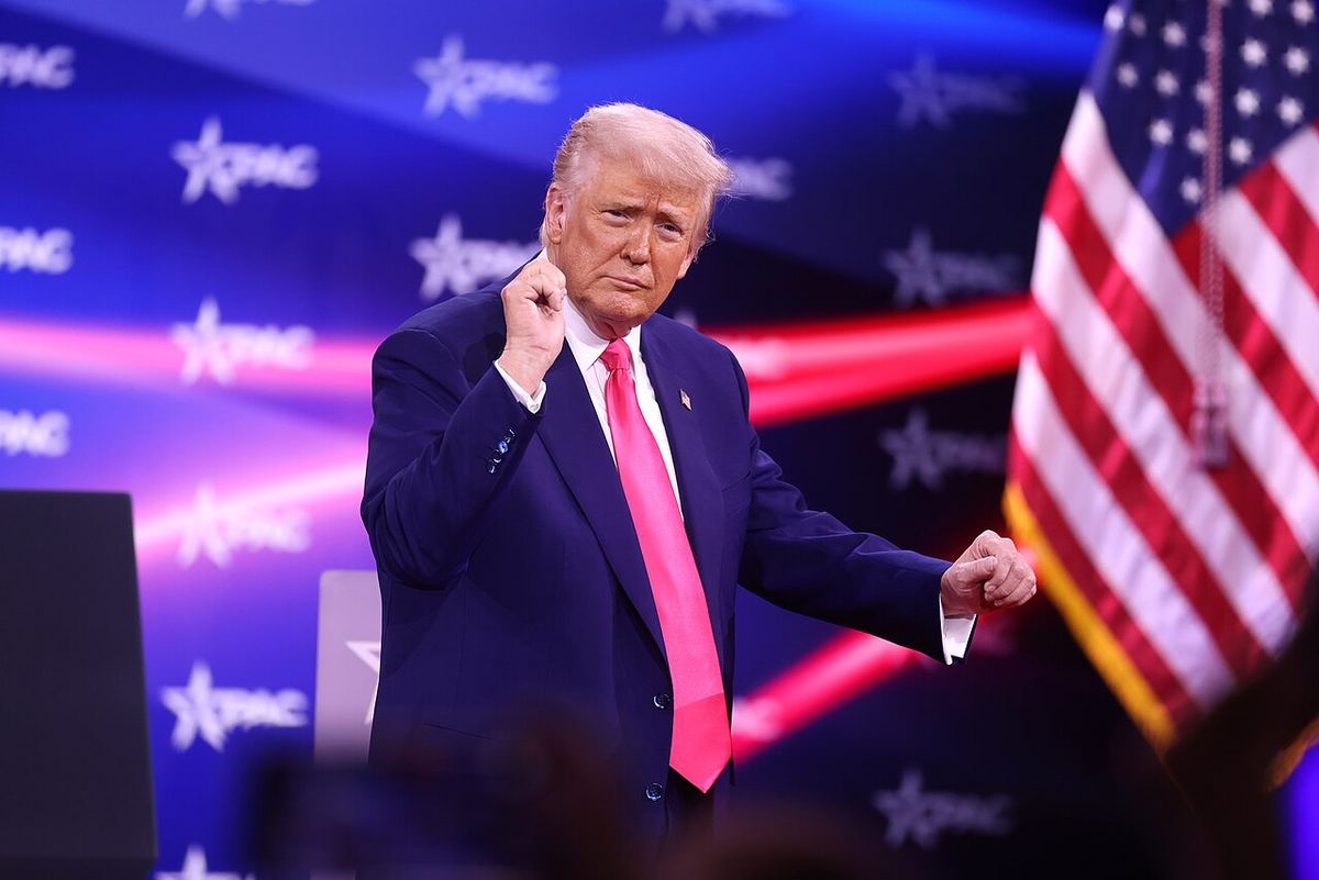 Donald Trump gesticulând în fața unui fundal albastru cu logo-uri CPAC, lângă un steag al SUA (fotografie de arhivă).