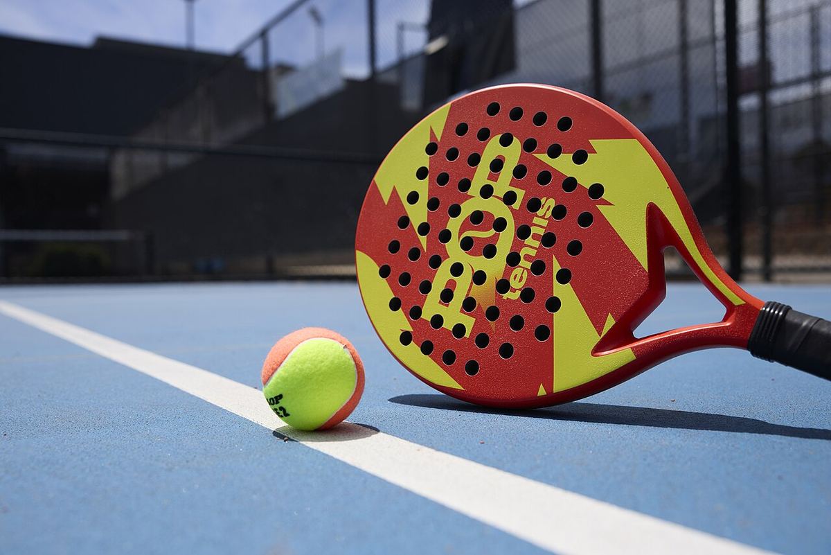 Paletă de padel roșie și galbenă și o minge pe un teren albastru, lângă o linie albă.