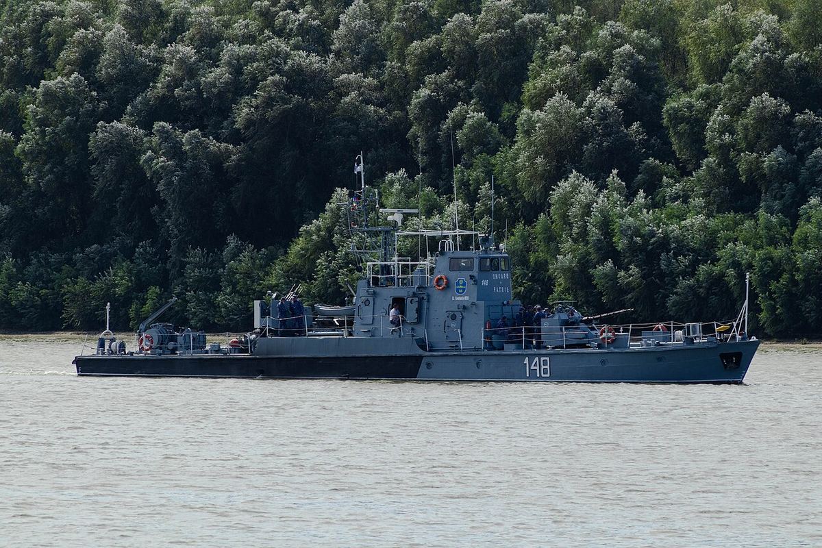 Navă de patrulare cenușie, cu numărul 148 pe corp, navigând pe o apă liniștită în fața unei păduri verzi.