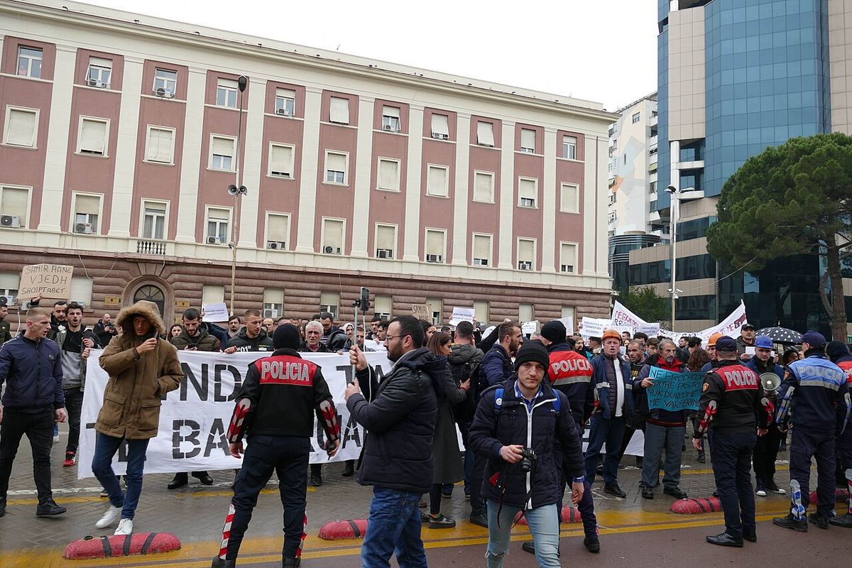 Manifestanți și polițiști în uniformă pe o stradă din Tirana, Albania, cu un banner alb în fundal.