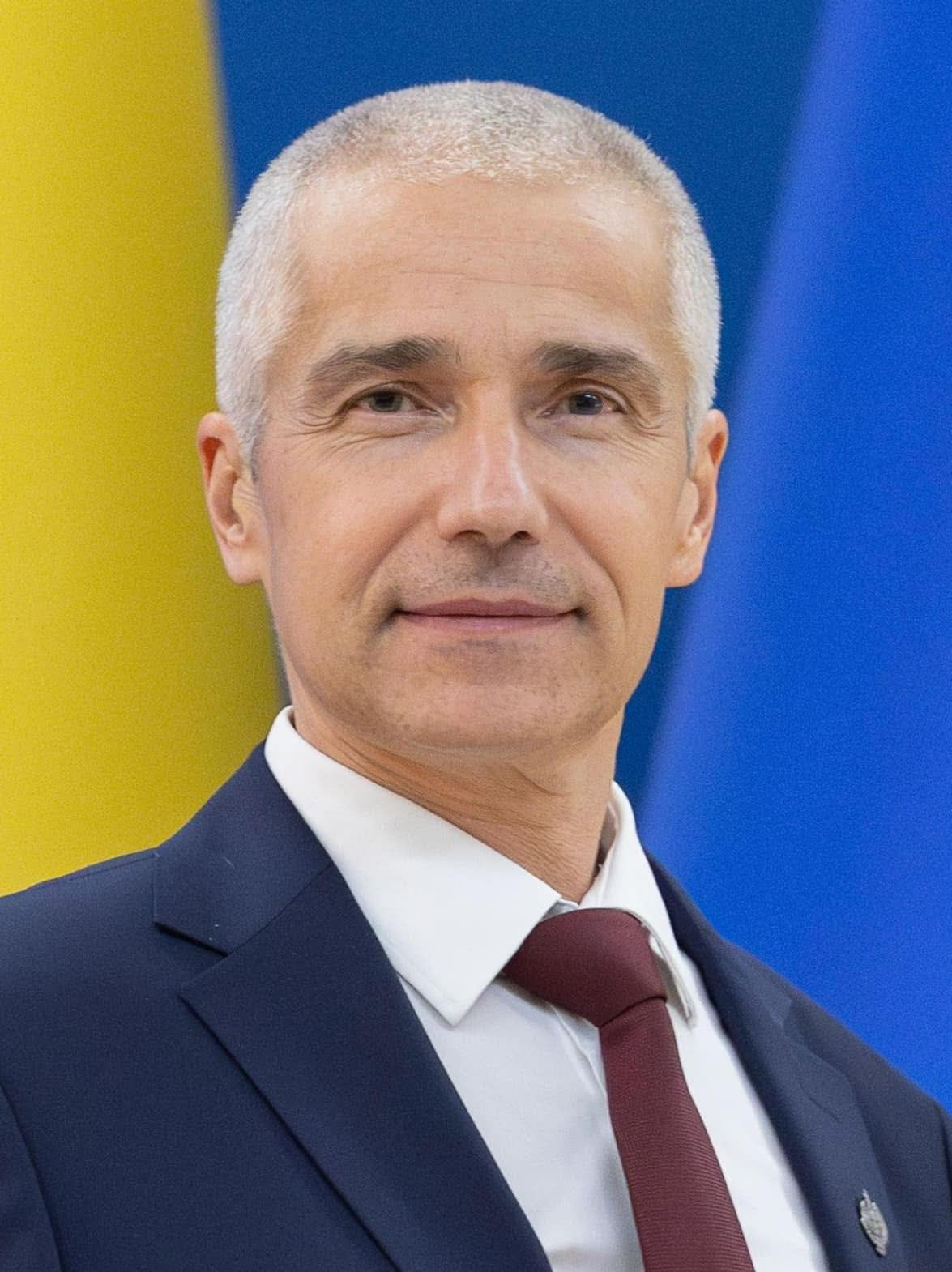Portretul lui Radu Marinescu, ministru al Justiției, în costum bleumarin și cravată bordo, în fața unui fundal galben și albastru.