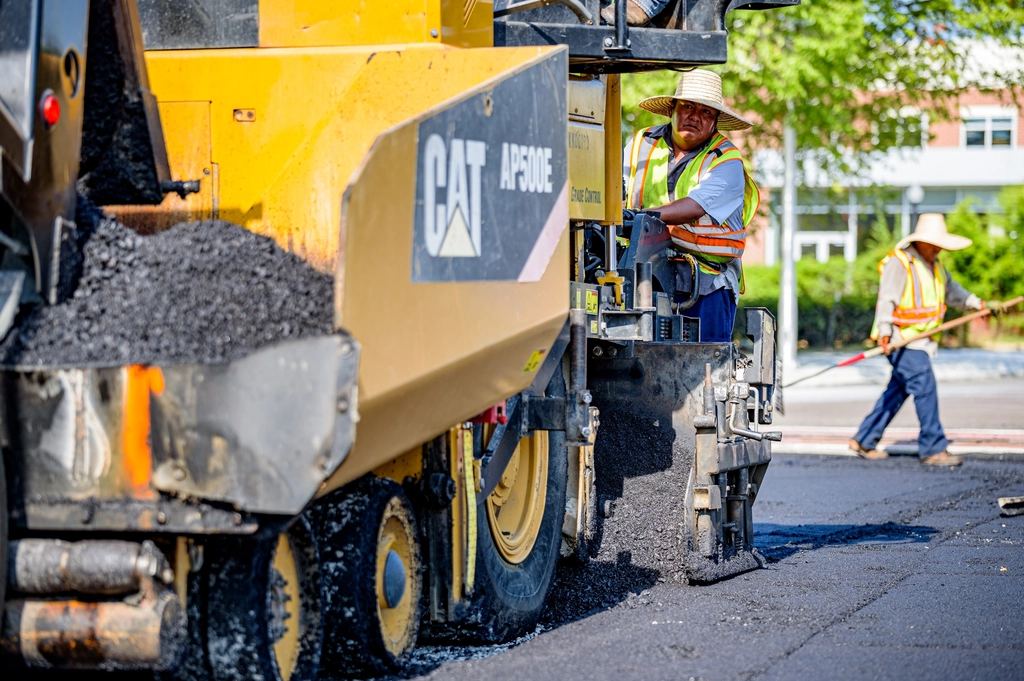 Utilaj de asfaltare Caterpillar așterne asfalt proaspăt, lângă muncitori în veste reflectorizante, pe o stradă.